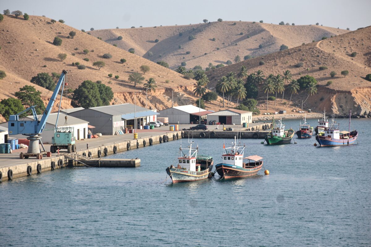 Un port de commerce en rade abritée, quais et grues modestes, boutres et bateaux de pêche au mouillage, collines sèches autour.