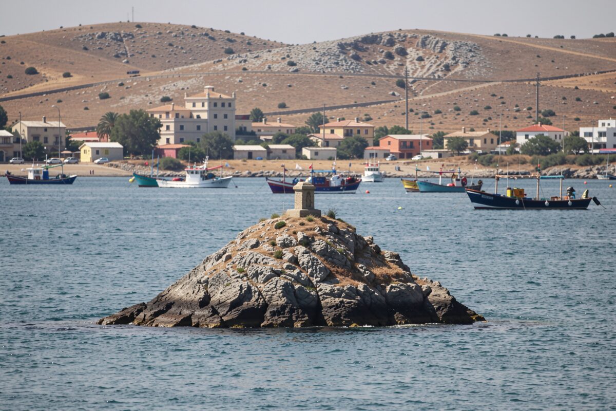 Un îlot rocheux conique au milieu d'une grande baie, eau verte, bateaux de pêche au mouillage, ville basse et collines en arrière-plan.
